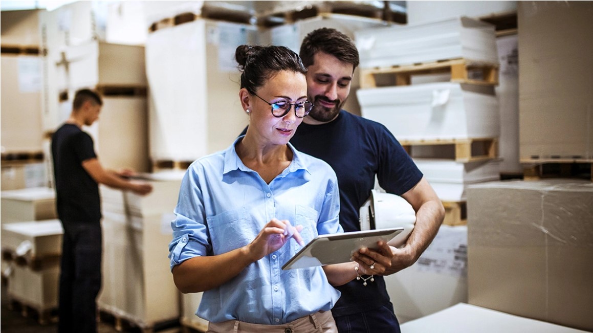 Woman reviewing information on a tablet in a warehouse with boxes and other workers around her