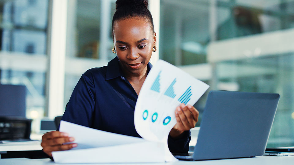 Accountant reviewing printed financial reports and charts while working at a desk with a laptop.