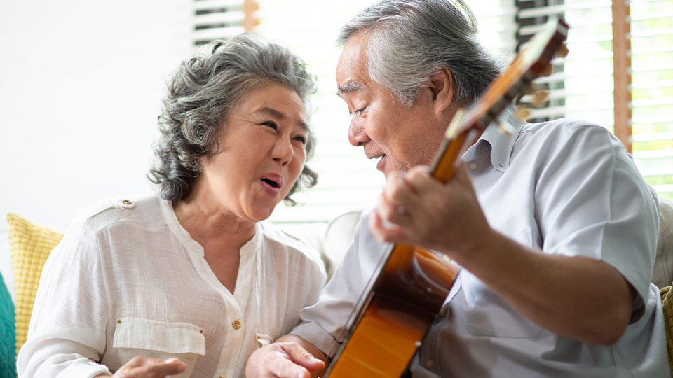A gray haired man sits holding a guitar next to a gray haired woman who is singing along