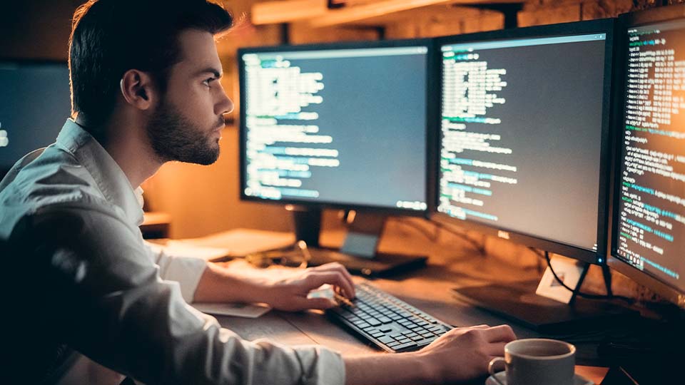 A bearded man sits in front of three computer monitors analyzing website back end code
