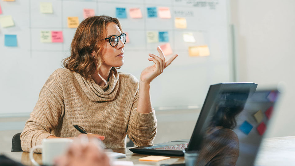 A woman looks engaged in a conference room with colorful sticky notes on a whiteboard in the background. Her laptop is open and she has a pen in her hand.