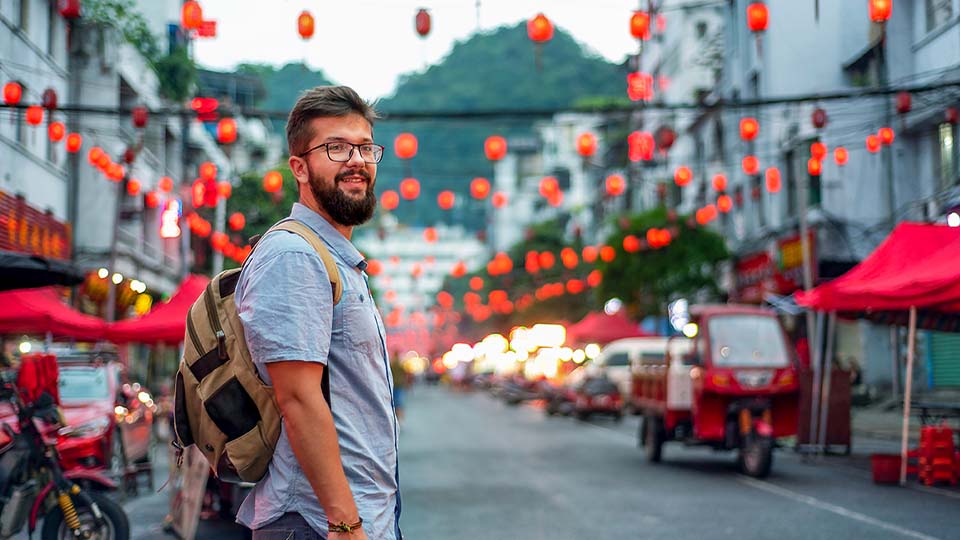 A bearded man with glasses and a backpack stands on a street with Chinese red lanterns hanging on wires above