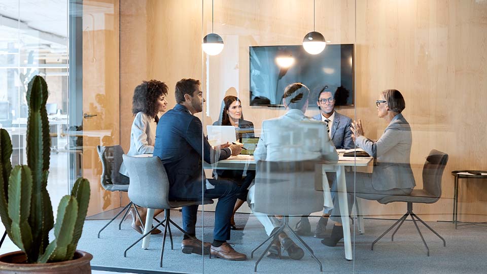 A diverse group of people sit around a conference room table, listening to a woman at the head of the table speak