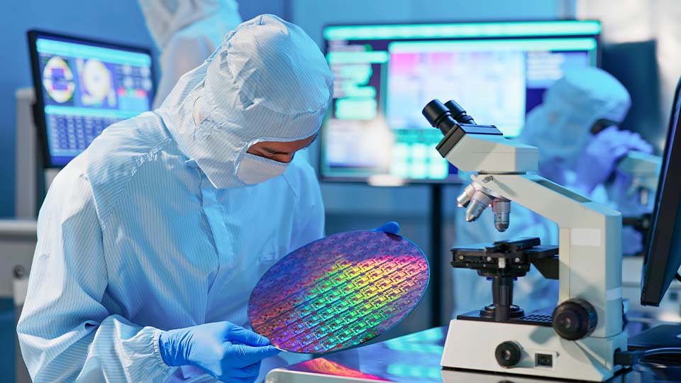 Chemical engineer in cleanroom gear inspects a silicon wafer in a high-tech lab with microscope and data monitors