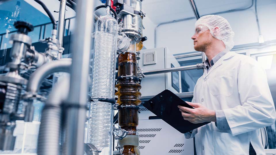 Chemical engineer in lab coat inspects industrial distillation equipment while holding a tablet in a high-tech processing facility