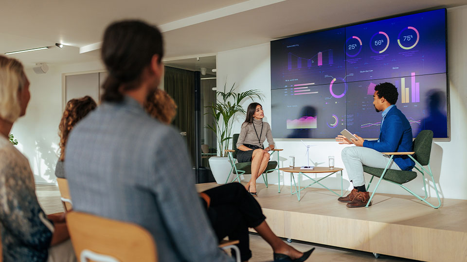 A seated audience listens as panelists discuss and gesture during a presentation with data visuals behind them