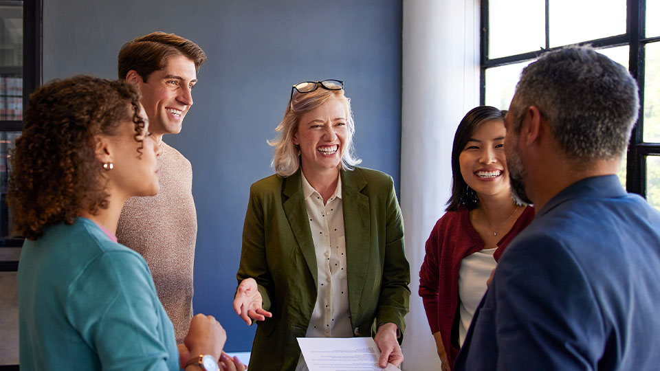 A group of five professionals in a modern office engaged in a collaborative discussion, sharing ideas and reviewing printed materials
