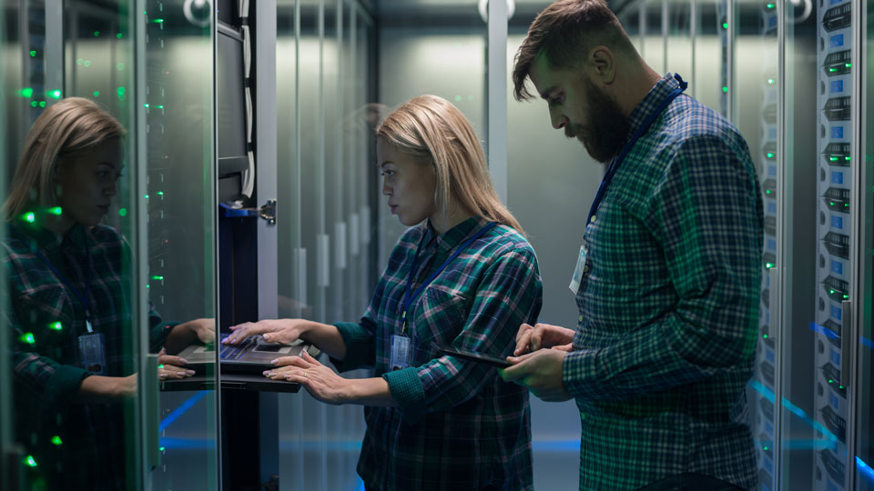 Two people working in a dimly lit data center with servers, with a woman typing on a keyboard and a man using a tablet