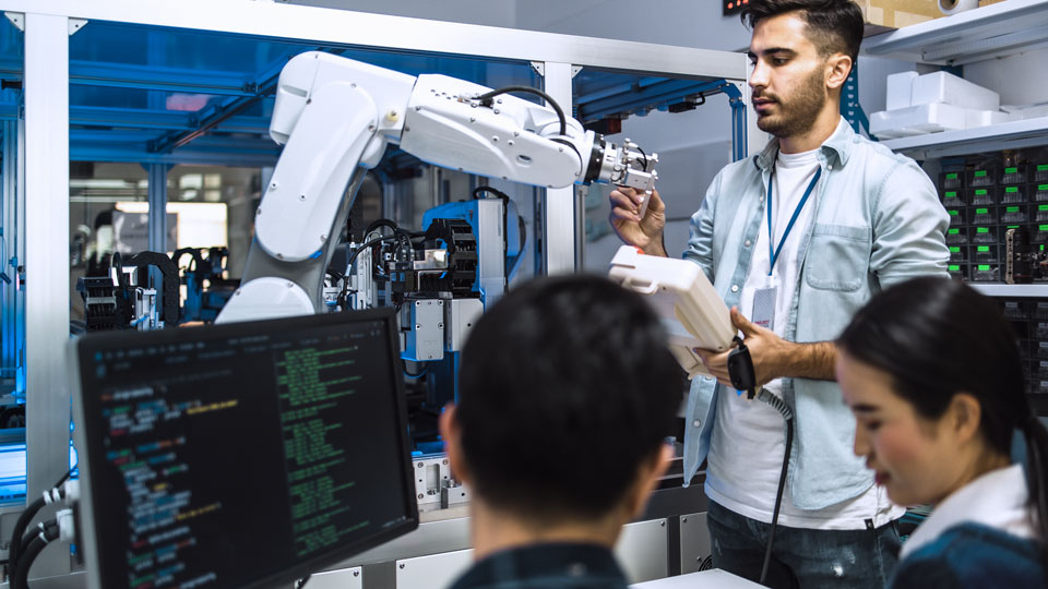 A man operates a robotic arm in a technology lab, with a computer displaying code in the foreground