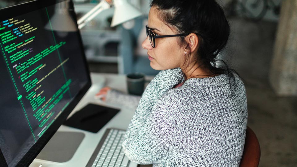 Woman in glasses and gray sweater reviewing code on her computer screen