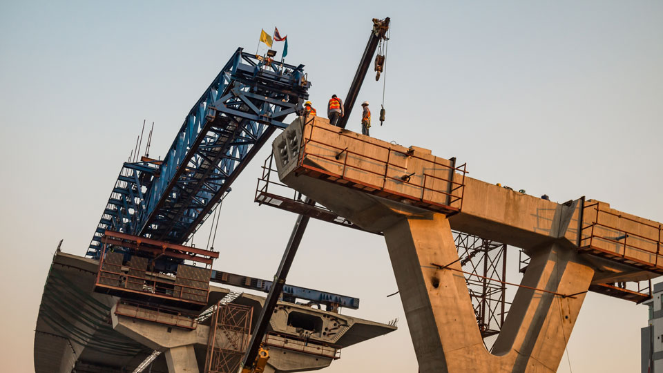 Workers at bridge construction site stand on a concrete pier with a blue truss structure and crane high above