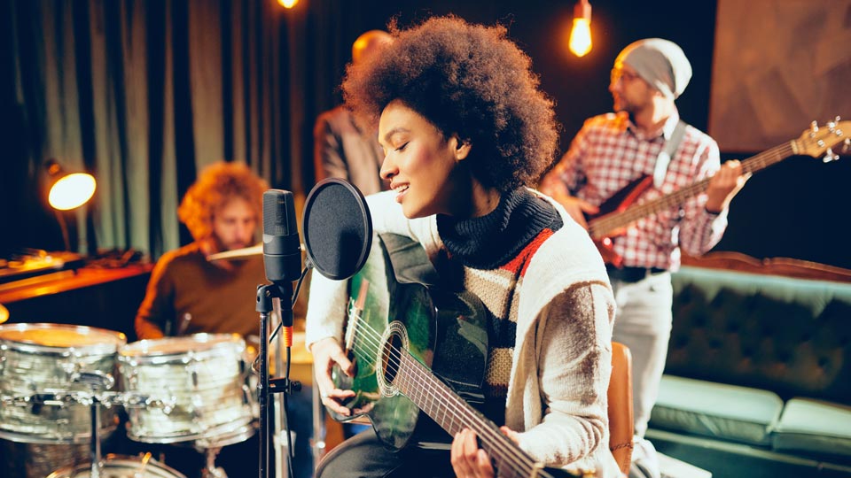 Woman holding a guitar and singing into microphone in studio setting with bandmates behind her