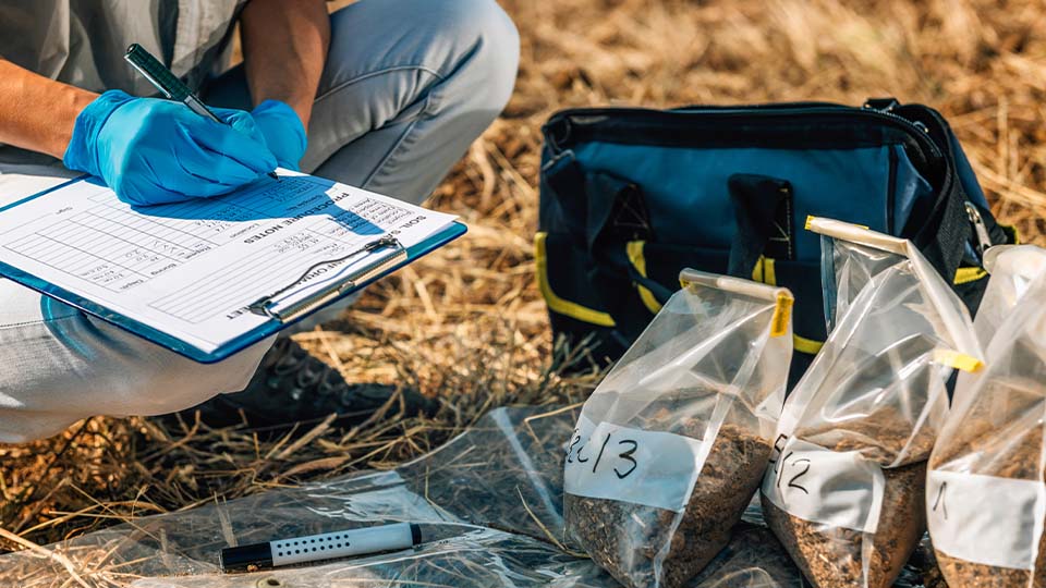 Person in blue gloves squats while writing on a clipboard in a field with soil samples and tools nearby