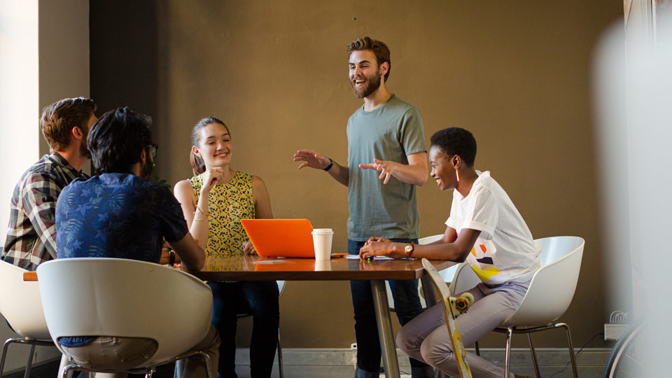 A group of five people having a discussion around a wooden table in a brightly lit room, with one person standing