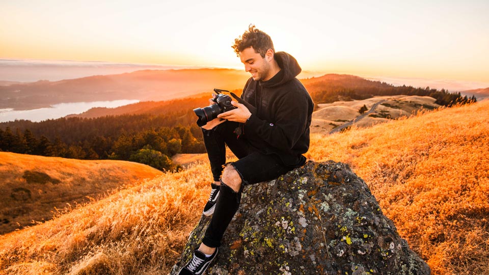 Young man sitting on hillside reviewing shot on DSLR camera with beautiful sunset behind him