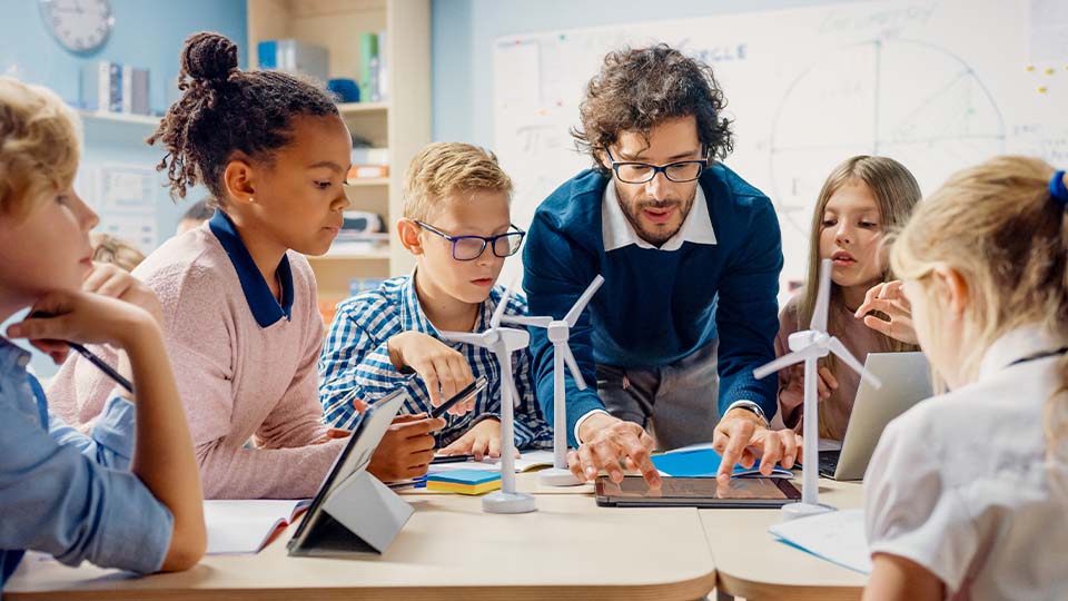 Elementary education teacher guiding students in a classroom STEM activity with wind turbine models and notebooks on the table
