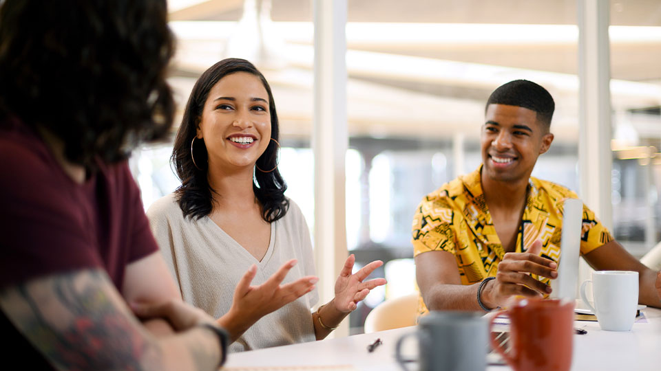 Woman speaking with hand gestures during a group discussion while others listen and respond around a table.