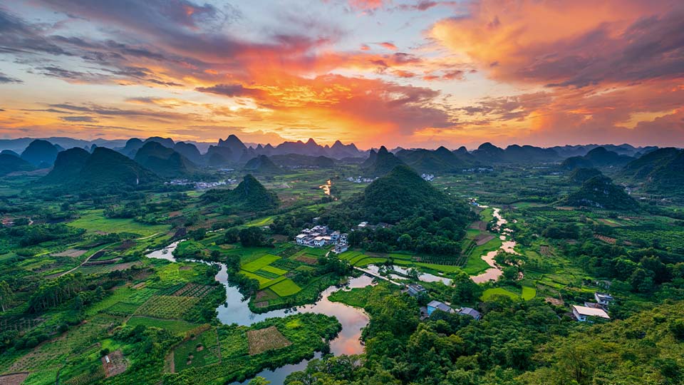 A picturesque valley with a river winding through green fields and karst mountains at sunset