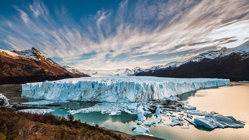 A blue glacier feeds into a small lake, surrounded by snow-capped mountains and a cloudy sky