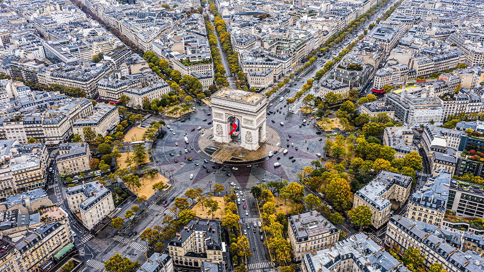 Aerial view of the Arc de Triomphe in Paris surrounded by a circular road and city buildings, with tree-lined streets and cars visible.