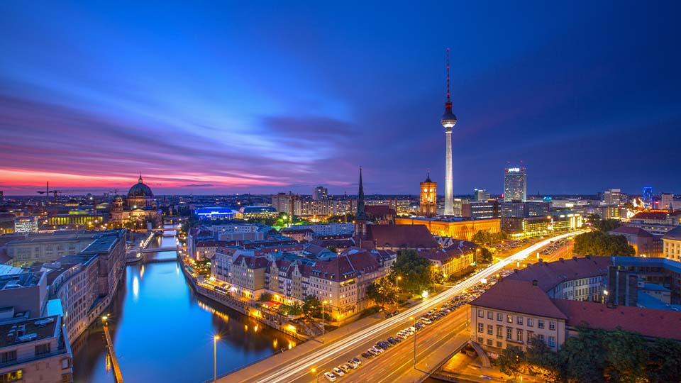 An aerial view of Berlin at dusk, featuring the TV Tower and river, with city lights reflecting on the water and a colorful sky.