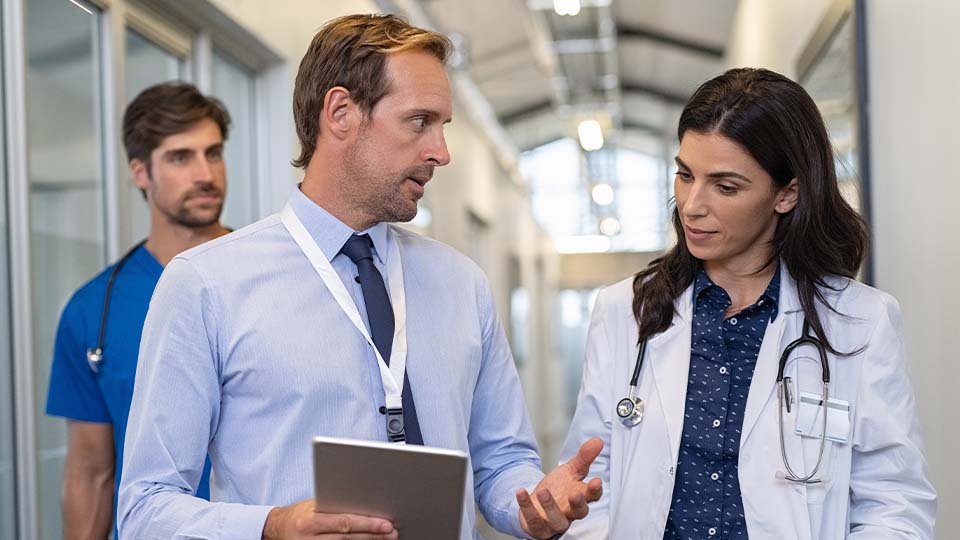 A medical administrator and a care provider walk and talk in a hallway as a nurse listens from behind