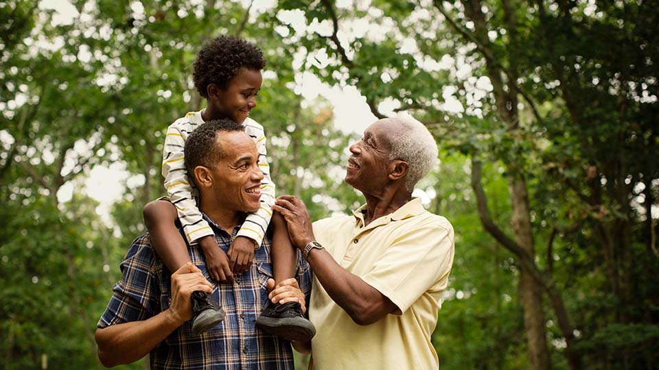 Three generations of a family enjoying time outdoors, with a man carrying a young child on his shoulders and an older man smiling warmly