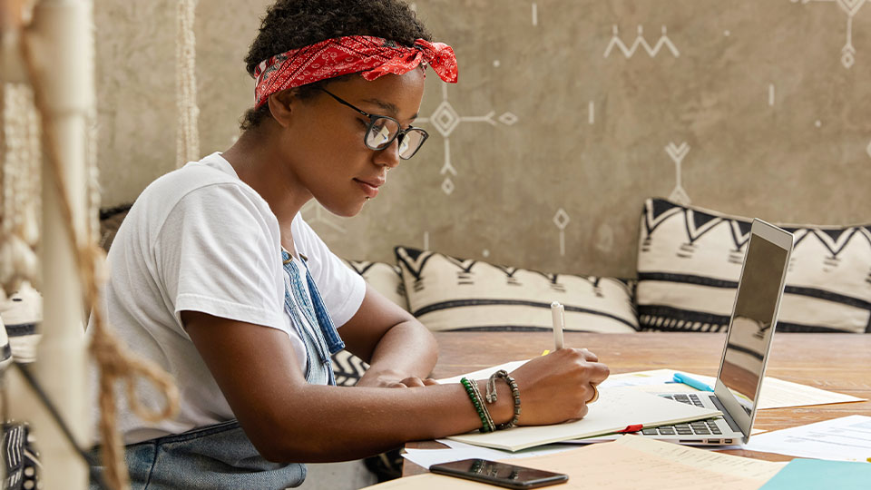 A young woman with glasses and a red headband writes in a notebook at a wooden table with a laptop open in front of her.