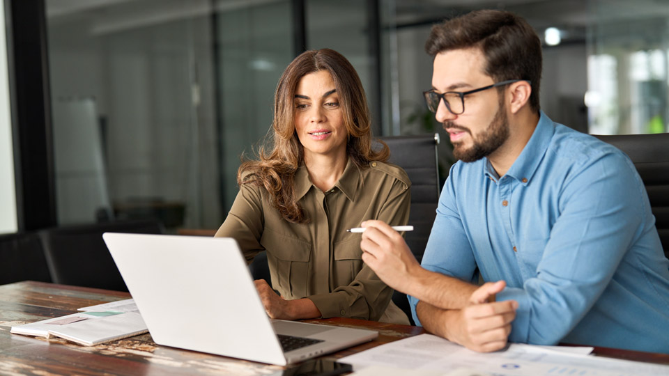 A man with glasses and a woman sitting at a table, looking at a laptop. The man holds a pen in his hand