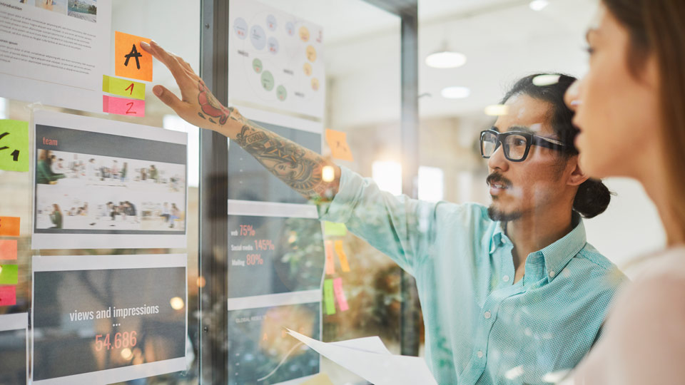 Man in glasses with forearm tattoos gesturing toward printouts of site design and post-its taped to glass wall