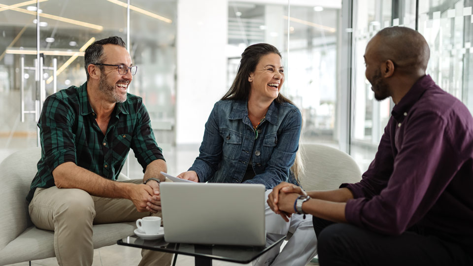 Three people sitting and laughing in a modern office, with a laptop and a coffee cup on the table