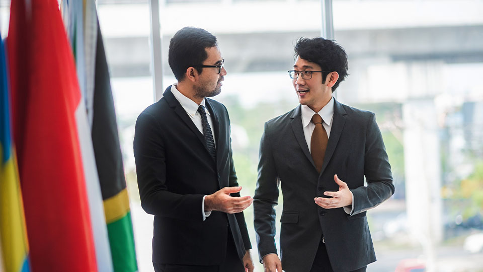 Two men in suits having a conversation in a brightly lit room with international flags in the foreground