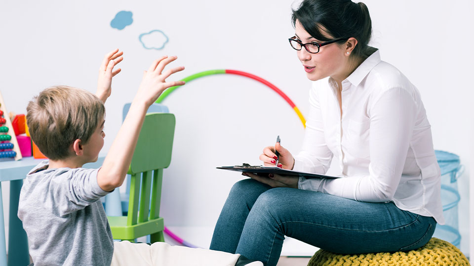 Woman taking notes on clipboard while talking to a young child with toys in background