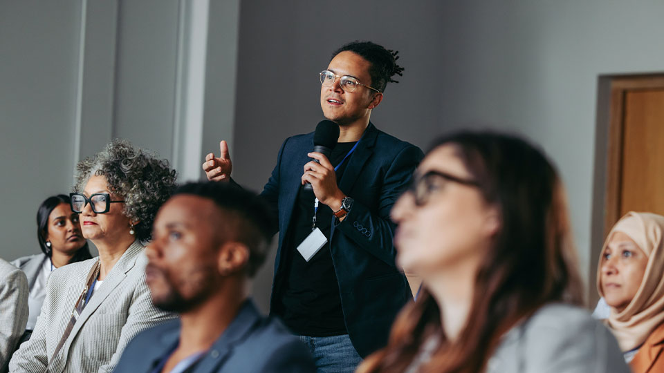 A person in the audience of a public event holds a microphone and speaks while facing the front of a room as attentive audience members listen.