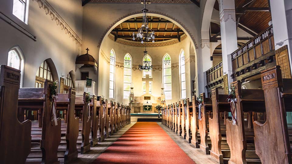 The interior of a church with wooden pews, arched windows, a red carpet aisle and sunlight streaming toward the altar at the front.
