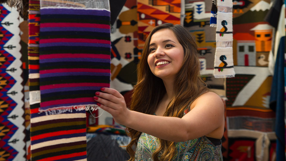 A smiling woman with long brown hair holds up a colorful textile, surrounded by various patterned fabrics in an outdoor market.
