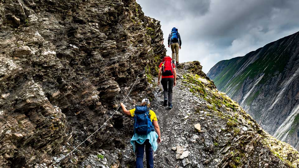 Three hikers walk along a narrow, rocky mountain path under overcast skies