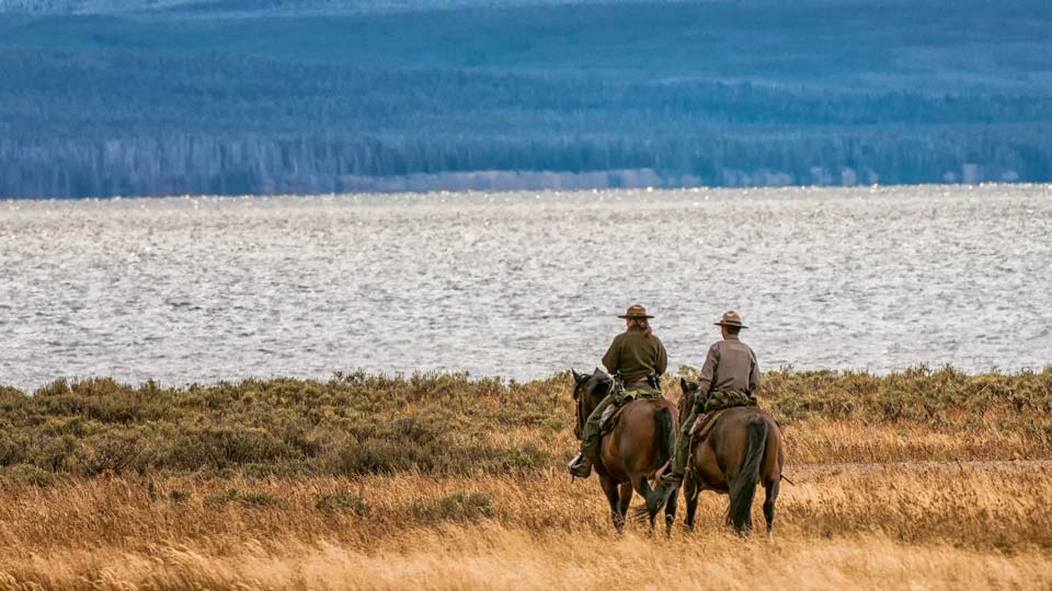 Two park rangers on horseback ride through tall grass, heading towards a large body of water with distant forested hills in the background