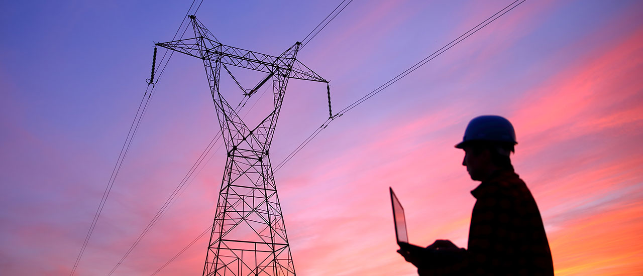 A power repair technician stands in front of a large power transmission tower at sunrise