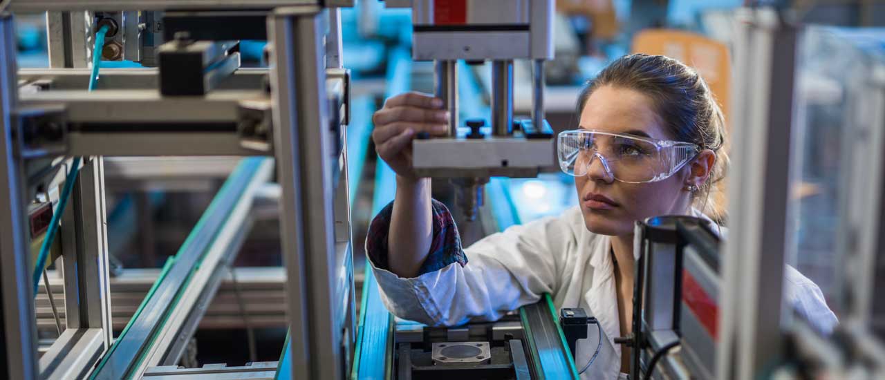 A woman wearing safety glasses and a white lab coat inspects a piece of machinery on a factory line