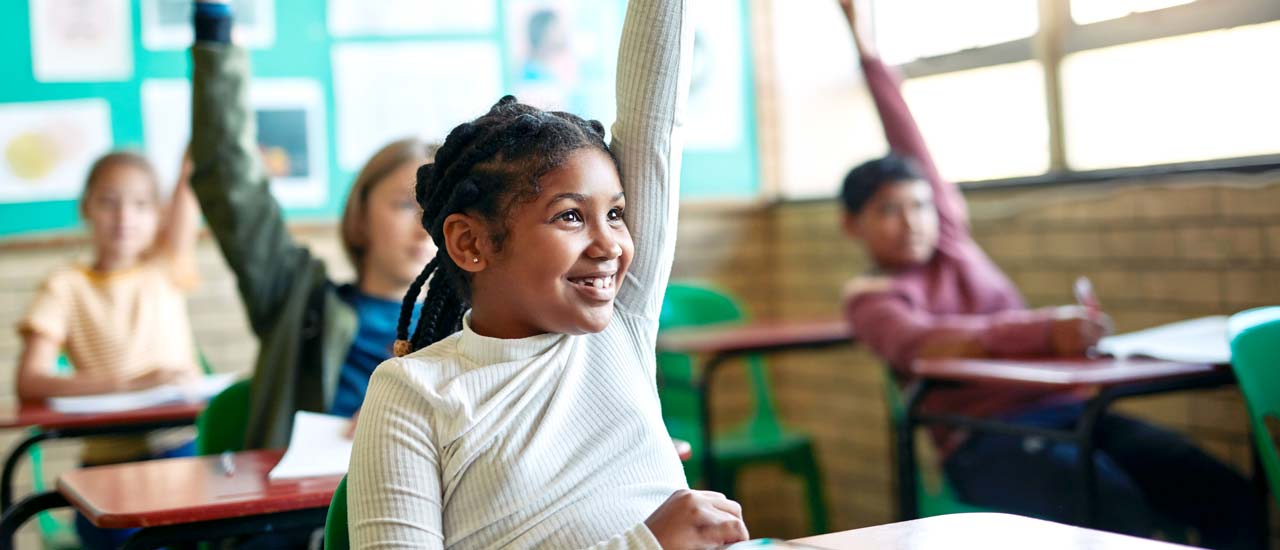 A young girl with her hair in box braids smiles as she raises her hand while her classmates raise hands in the background