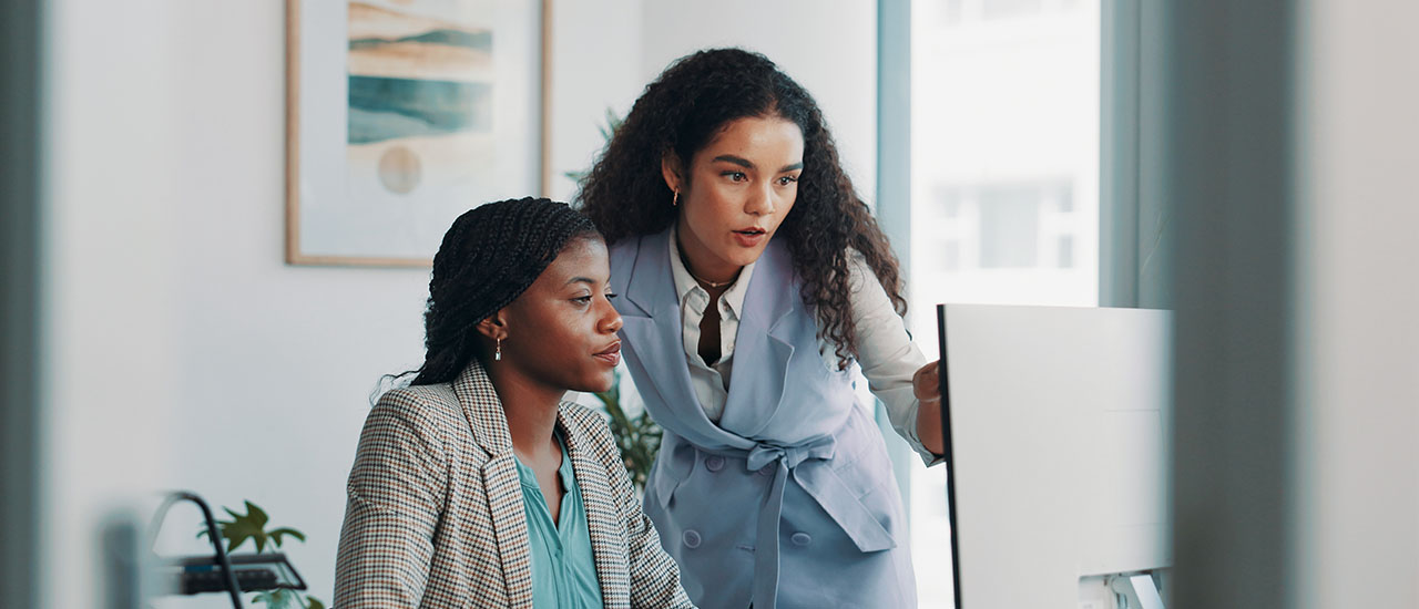 Colleague leans over a desk to review information on a computer screen with a seated IT professional.
