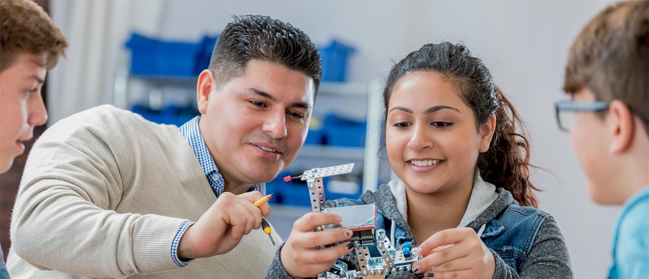 A male teacher works with a group of students on building a rudimentary hand-held robot. A young girl holds the device.