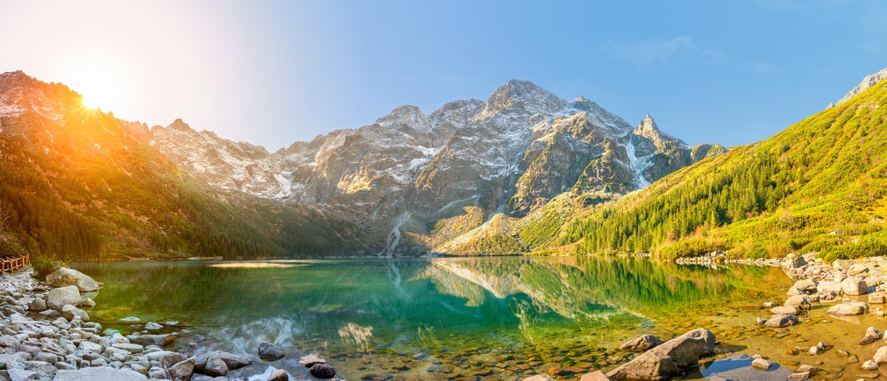 In a panoramic view of Morskie Oko, a lake in the Polish Tatra Mountains, the sun crests over a rocky peak behind the lake