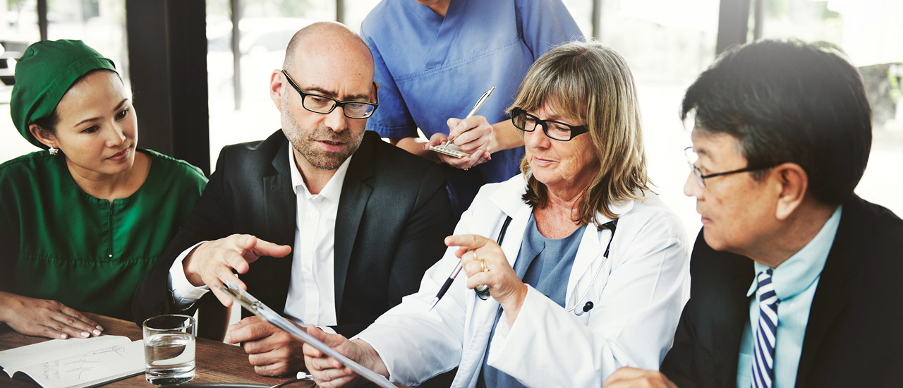 Four people seated at a table and one person standing behind them. Everyone is looking at a clipboard being held by a medical professional.