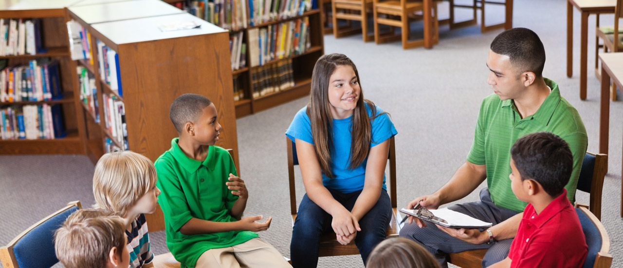 A school counselor holds a clipboard while he sits with a group of diverse children in a circle
