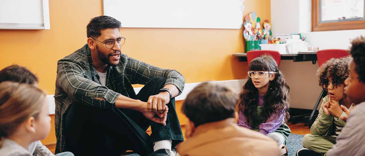 A male teacher sits on the floor with young students in a circle, engaging them in discussion in a colorful classroom setting