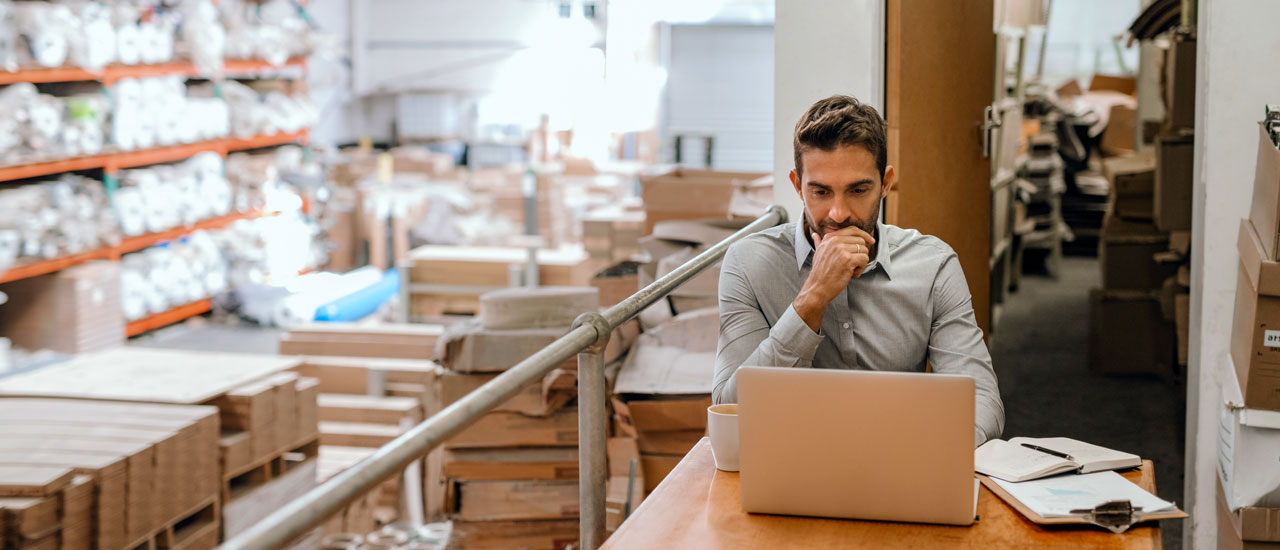 Man sitting at a table with an open laptop and stacks of shipping supplies behind him
