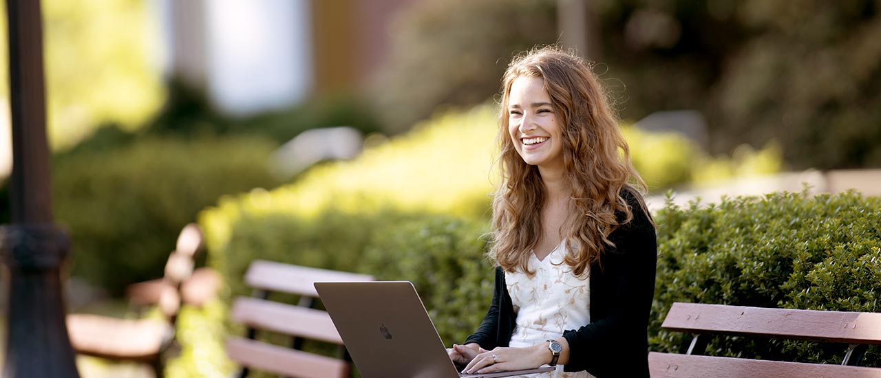 Wide angle of girl with laptop sitting on outdoor bench smiling in the sunshine