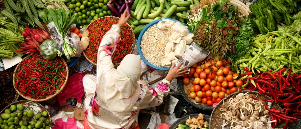 A woman in a white hijab reaches over large baskets full of brightly colored vegetables and fresh produce